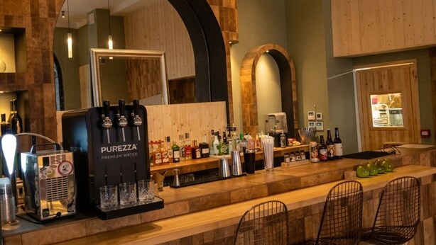 A bar area with a PURREZZA water dispenser, bar stools, and a wooden wall with a large mirror.