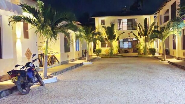 A building with a courtyard, palm trees, and a motorcycle parked outside. - Simba Apartments Diani  (Ukunda)