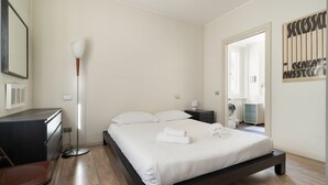 Ein Schlafzimmer mit Bett, Nachttisch, Lampe, Stuhl und Blick ins Badezimmer.