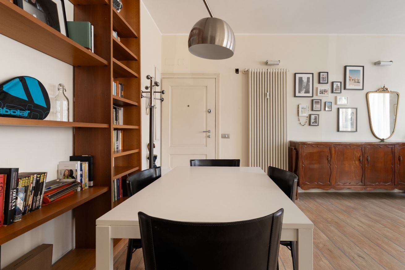 A dining area with a white table, black chairs, a wooden bookshelf, and a radiator.