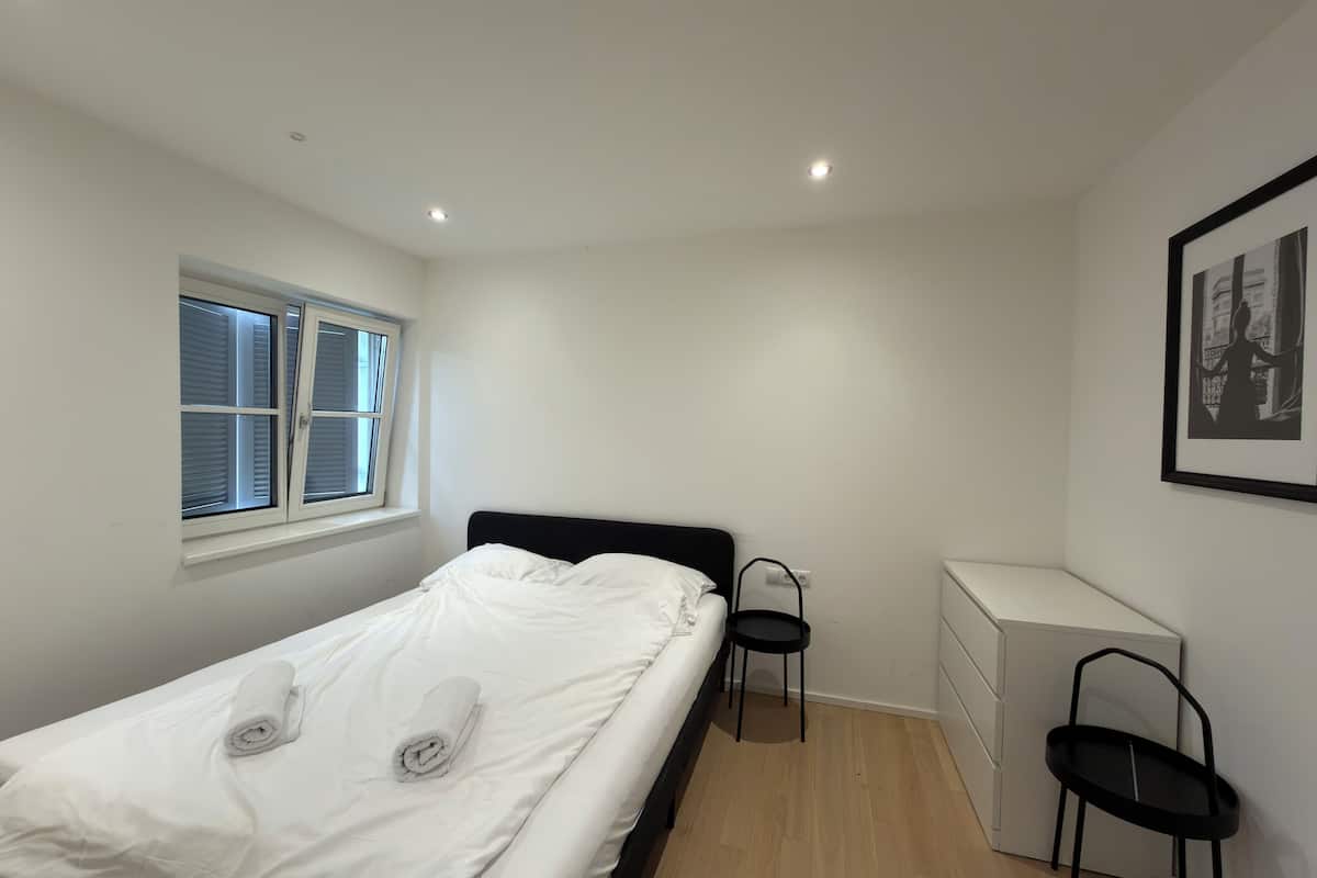 Une chambre à coucher avec un lit, une fenêtre, une chaise, un bureau et un tableau encadré au mur.