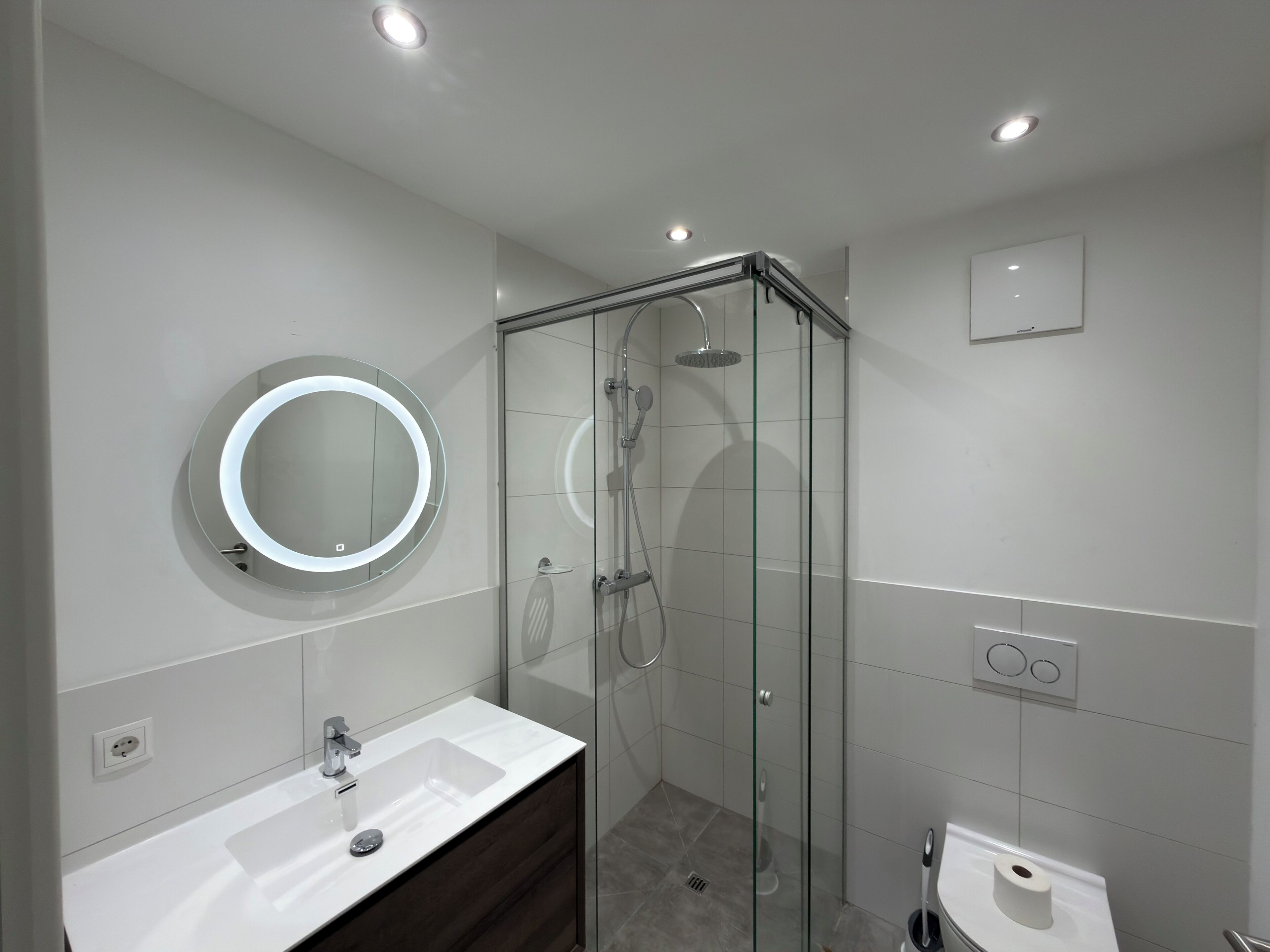 Een moderne badkamer met een douchecabine van glas, een ronde spiegel met verlichting, een witte wastafel en een toilet.