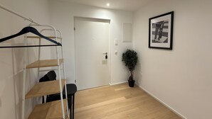 Una habitación con una puerta blanca, una obra de arte enmarcada en la pared, una planta en maceta, suelo de madera y un perchero con perchas.