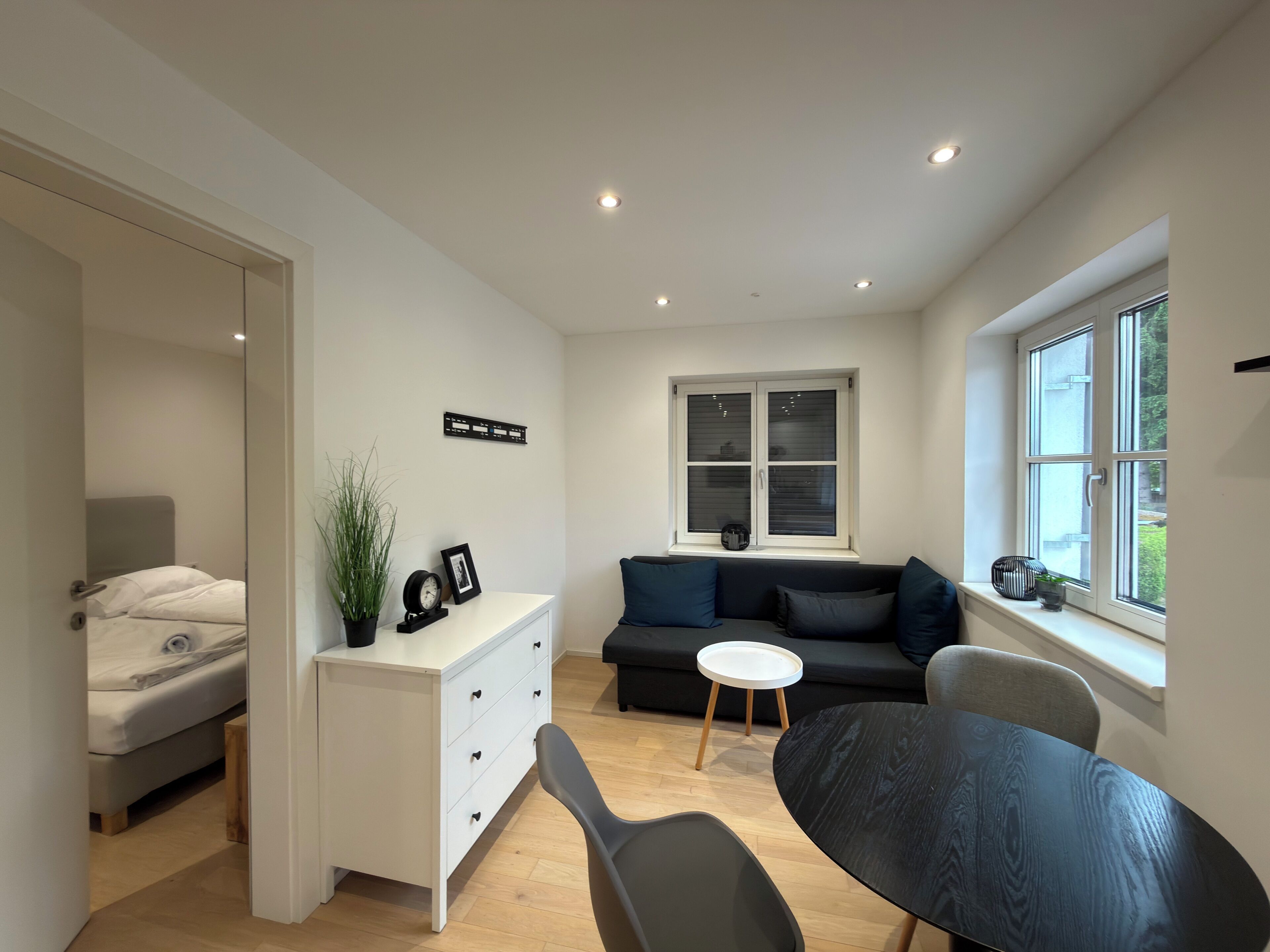 A modern, compact living space with a bed, a sofa, a small table, and a window with blinds.