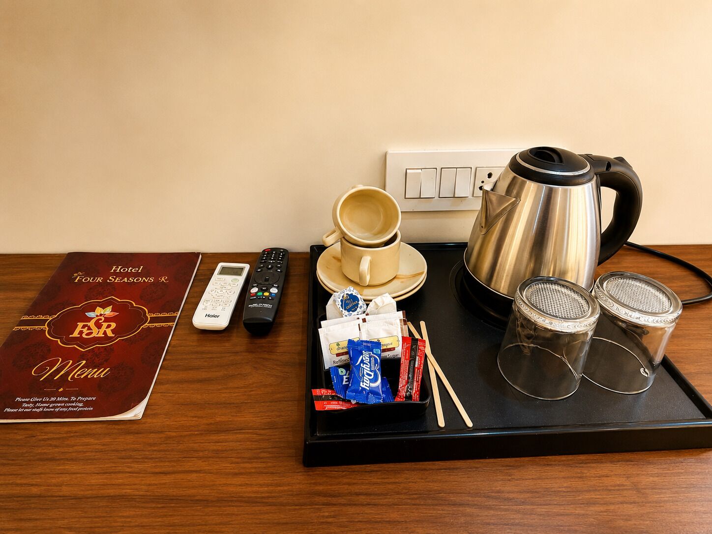 Un plateau contenant un menu d’hôtel, une télécommande, une cafetière, deux tasses, une bouilloire, des sachets de thé et des verres.