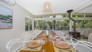 A dining area with a long table set for a meal, large windows, and a wood-burning stove. - Pet friendly,pool + stroll to beach and cafe (PEARL BEACH)