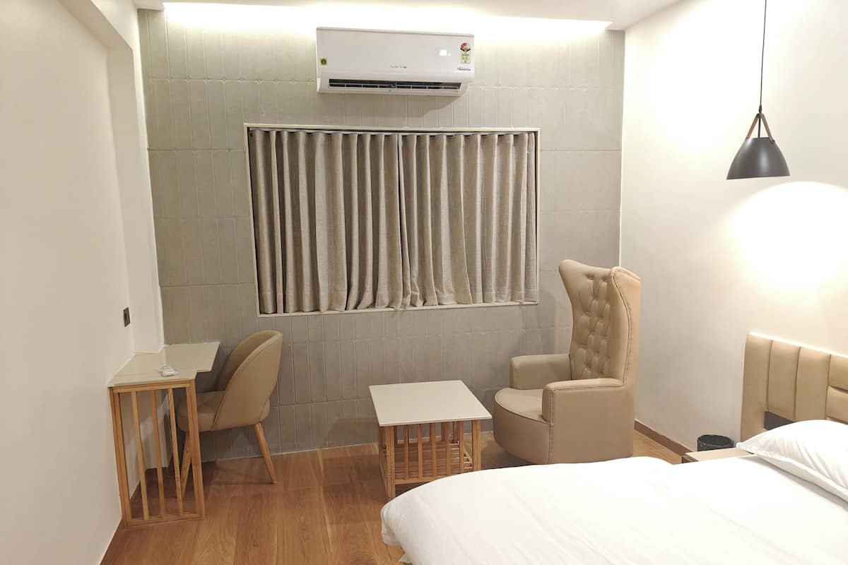 A hotel room with a bed, a desk with a chair, a small table, a cushioned armchair, a window with curtains, an air conditioning unit, and a hanging light fixture.