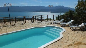 Una piscina con solárium de piedra, sillas de descanso y vistas a un lago y montañas.