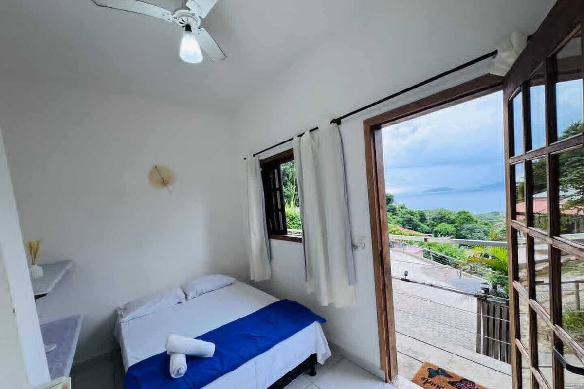 Ein Schlafzimmer mit Bett, Deckenventilator, Fenster mit Vorhängen und Blick ins Freie.