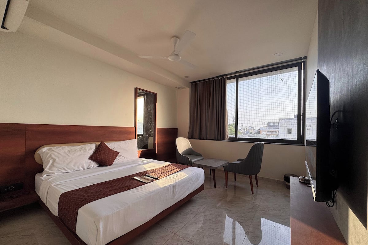 A hotel room with a large bed, a desk, a chair, a TV, and a window with a city view.