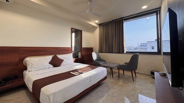 Quarto de hotel com cama, escrivaninha, cadeira, televisão, ventilador de teto e janela com vista para a cidade.