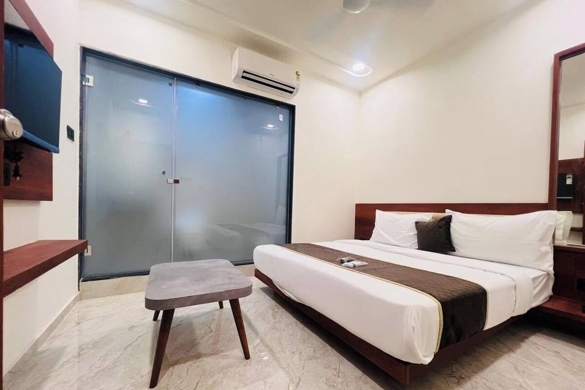 A hotel room with a bed, a small table, a wardrobe, and an air conditioning unit.