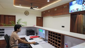 Um homem trabalhando em uma mesa com um computador, impressora e uma televisão fixada na parede.