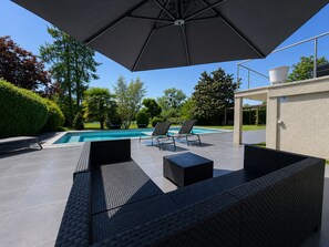 Un espace aménagé au bord de la piscine, comprenant un coin salon couvert, une piscine et une végétation luxuriante.