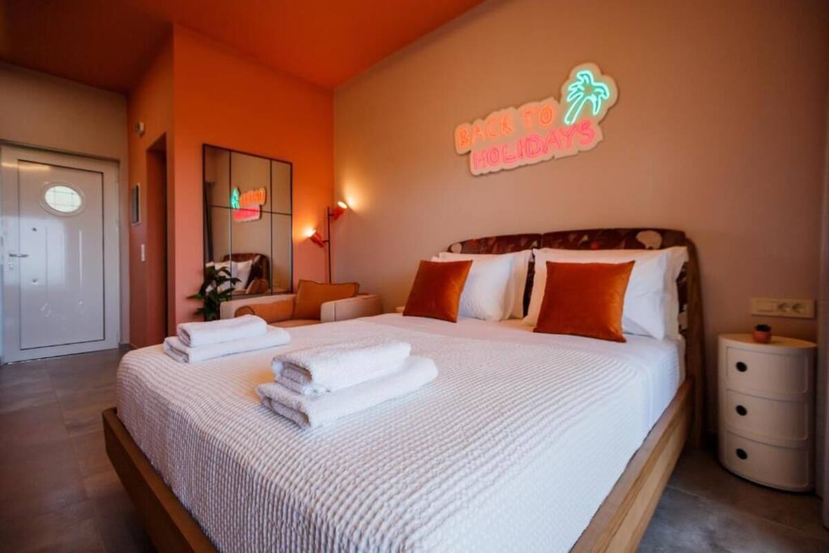 A hotel room with a bed, a nightstand, a mirror, and a neon sign on the wall.