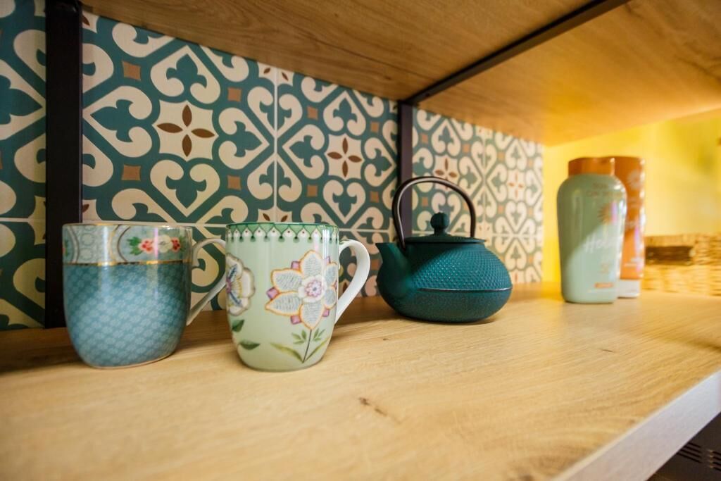 A shelf with a patterned tile wall, a blue mug, a floral mug, and a teal kettle.