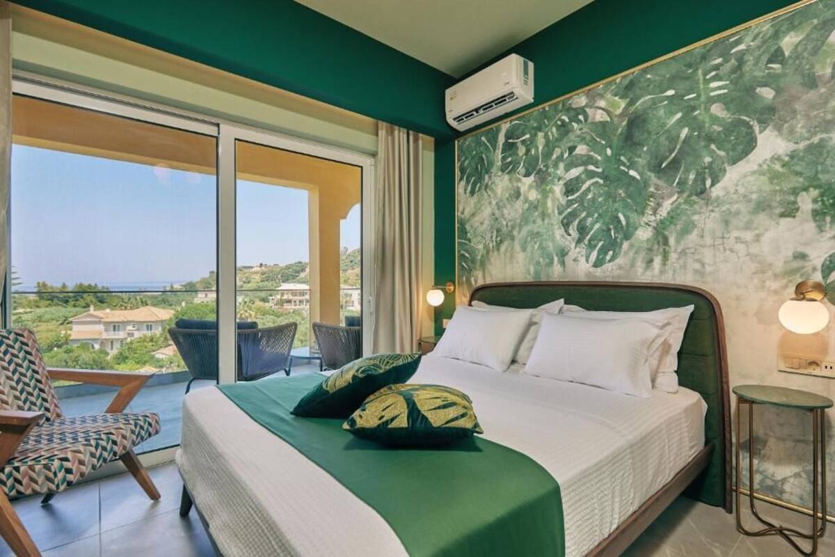 A hotel room with a large bed, a view of the sea, a balcony with a chair, and a tropical-themed wall painting.