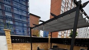 Eine Dachterrasse mit Gartenmöbeln, einer Pergola und Blick auf umliegende Gebäude.