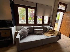 A grey sofa with cushions, a wooden floor, and a window with a view of a building and trees.