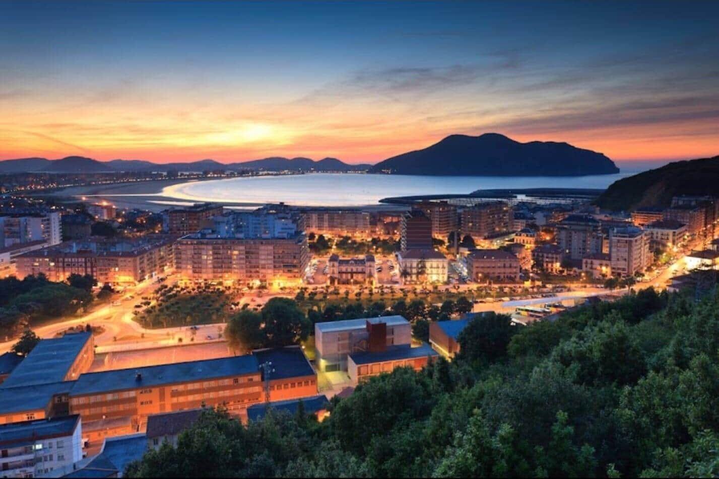 Un paisaje urbano costero al anochecer con edificios, carreteras y una masa de agua.