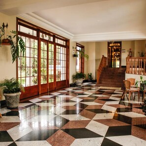 Un vestíbulo de hotel con piso a cuadros, grandes puertas de madera, una escalera y plantas en macetas.