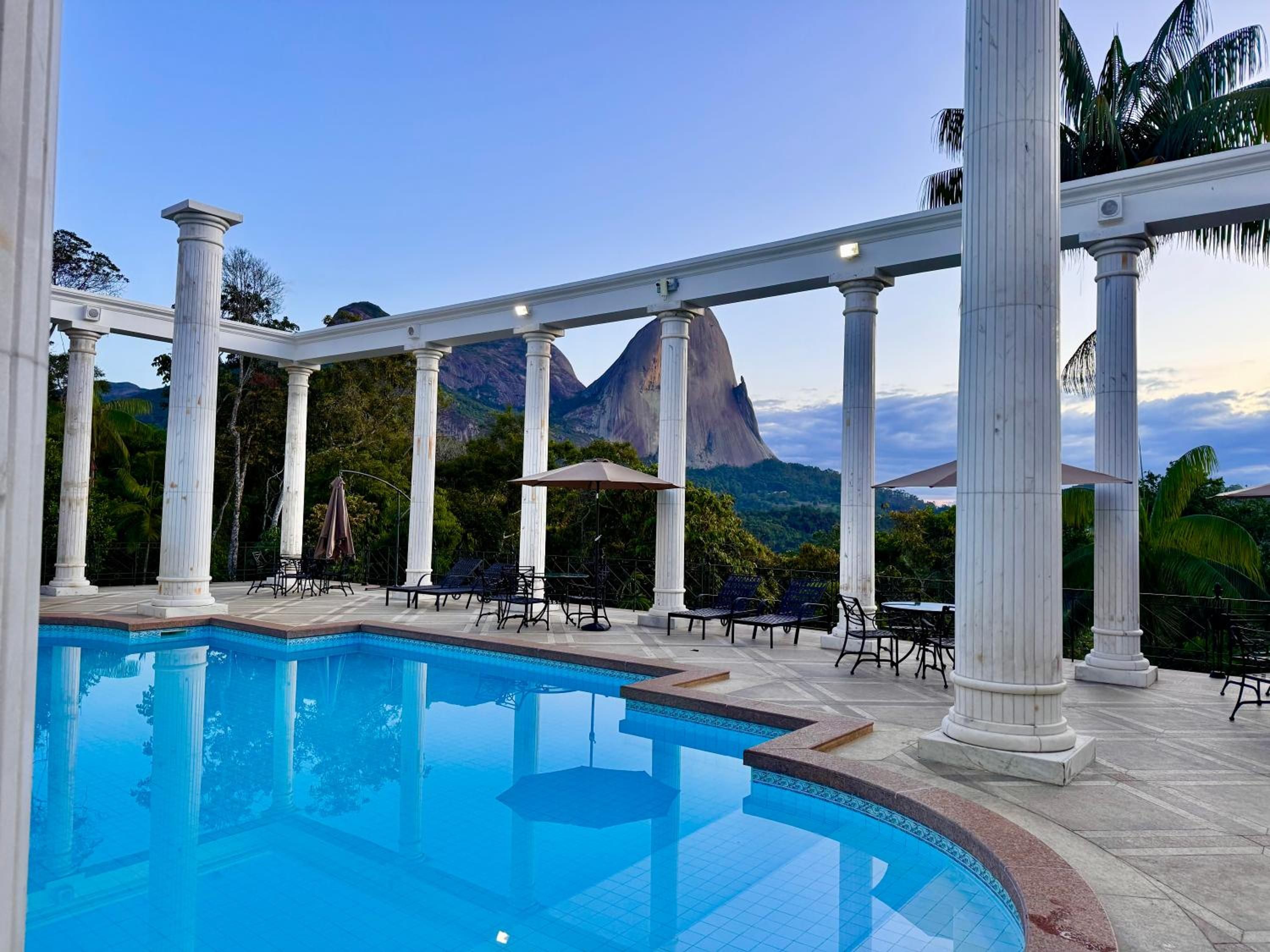 A pool area with columns, lounge chairs, and umbrellas, with a mountain view in the background.