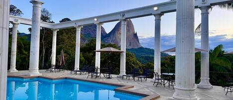 A pool area with columns, lounge chairs, and umbrellas, with a mountain view in the background.