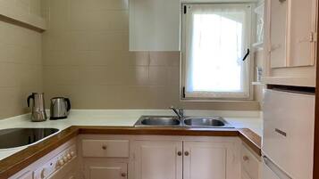 A kitchen with a double sink, a window with a white curtain, and a refrigerator.