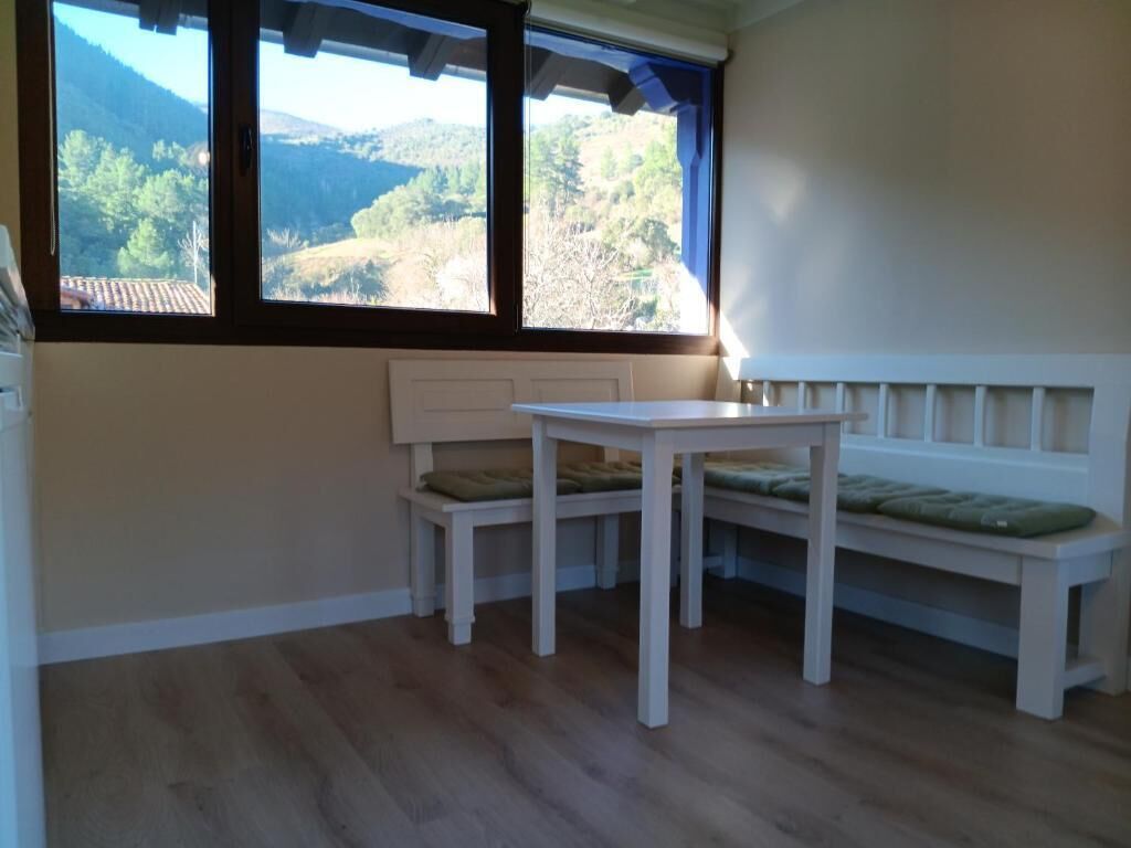 Una habitación con piso de madera, un banco blanco con cojines, una pequeña mesa y un ventanal con vistas a un paisaje montañoso.