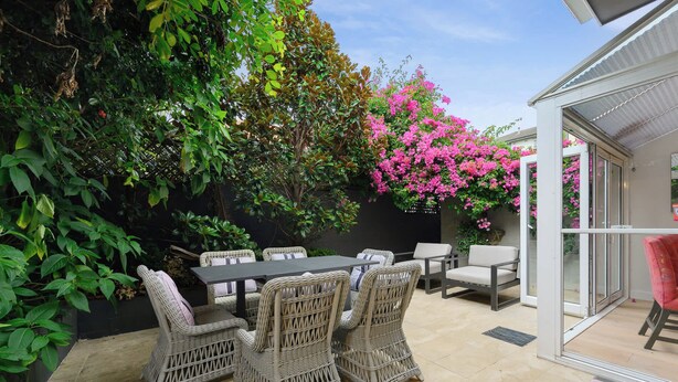 An outdoor patio with wicker furniture, a table, and chairs, surrounded by lush greenery and pink flowers. - Bright Townhome with Courtyard in Neutral Bay (Neutral Bay)