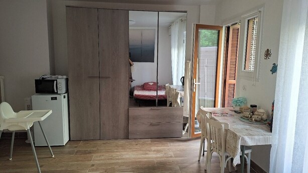 Quarto com uma mesa de jantar, cadeiras, uma geladeira branca e uma cama com colcha vermelha ao fundo.