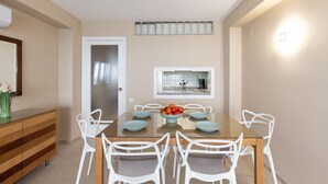 A dining area with a glass-top table, white chairs, a bowl of fruit, a window, and a kitchen visible through the window. - Beachfront | Spectacular sea views (La Pineda)