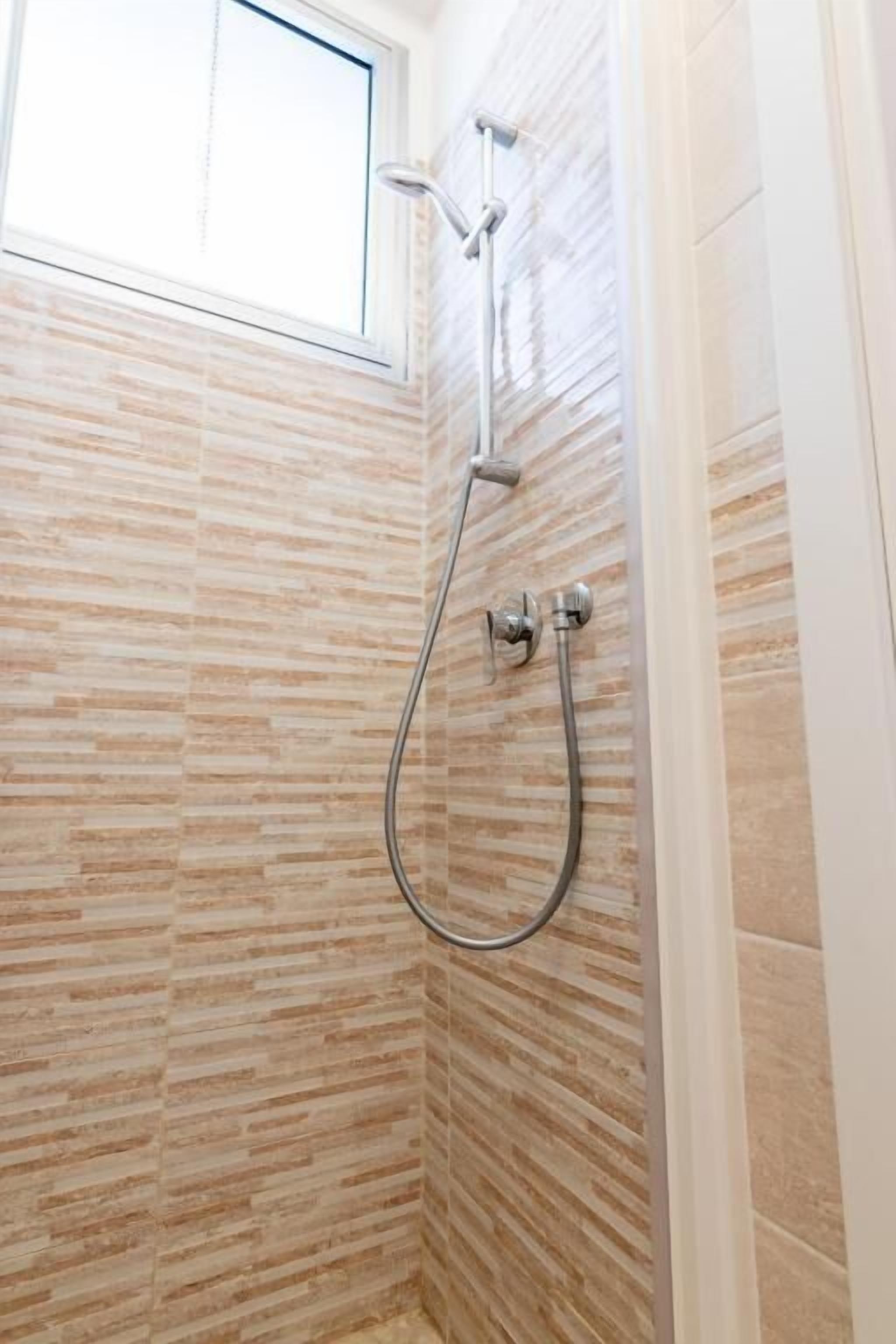 Ducha con cabezal de ducha de mano y grifo empotrado en la pared, paredes con azulejos y ventana.