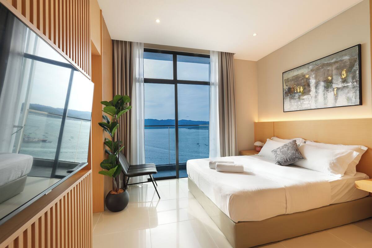 A modern hotel room with a large bed, a potted plant, a view of the sea, and a painting on the wall.