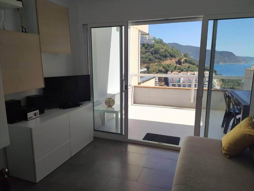 Ein modernes Wohnzimmer mit Fernseher, einer Couch und einem Balkon mit Blick auf Meer und Hügel.