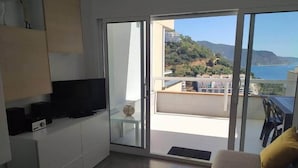 Ein modernes Wohnzimmer mit Fernseher, einer Couch und einem Balkon mit Blick auf Meer und Hügel.