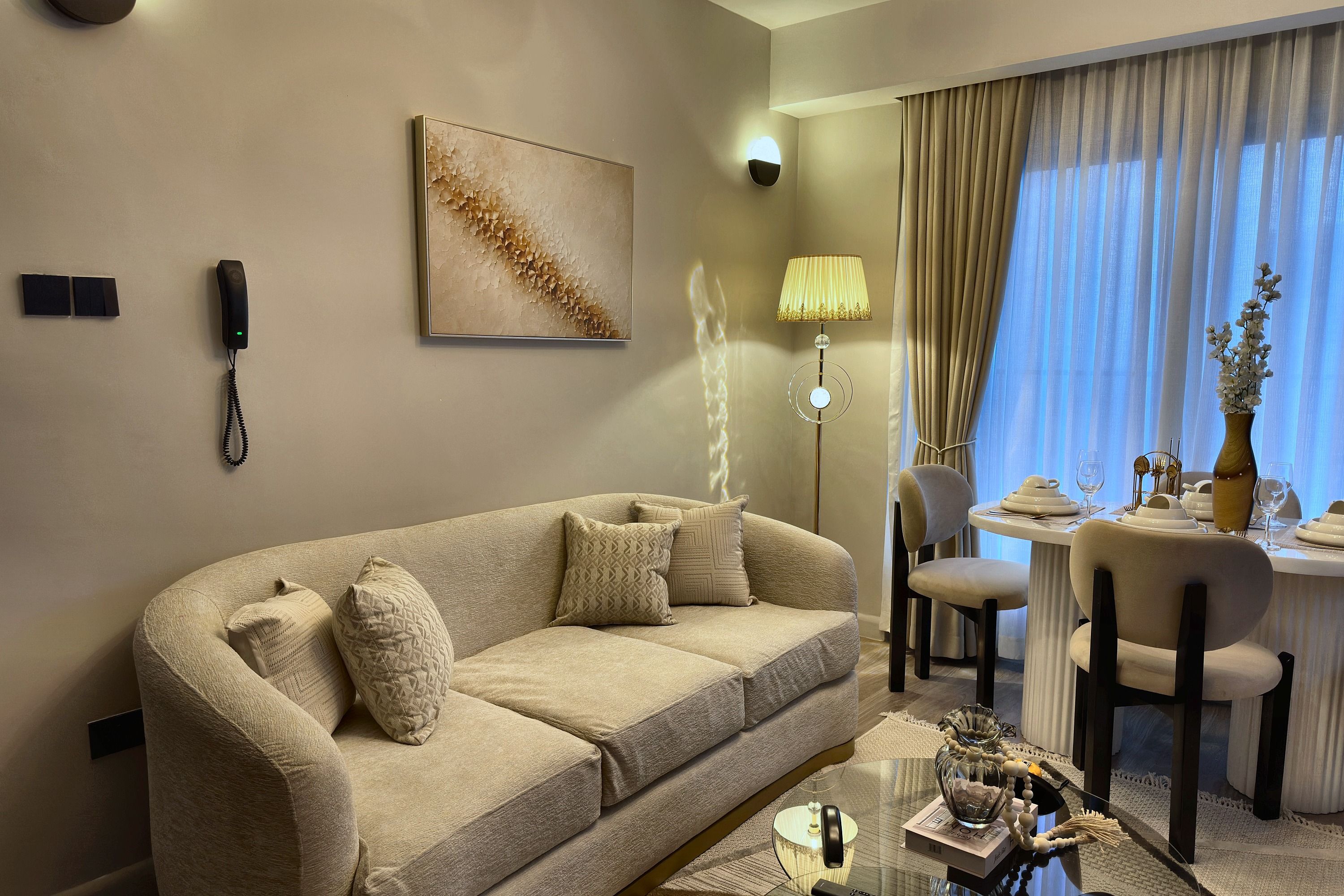 Habitación de hotel con un sofá color beige, un cuadro, un teléfono, una lámpara, una mesa de comedor para dos personas y un jarrón con flores.