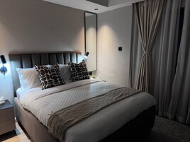 Quarto de hotel moderno com uma cama grande, cabeceira, abajures, mesa de cabeceira e um espelho amplo.