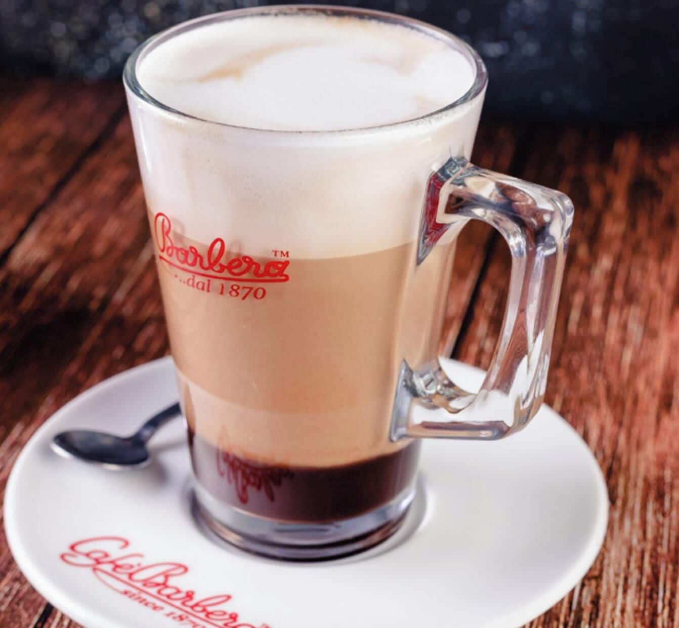 Une tasse en verre transparent, dotée d’une anse, contenant une boisson à plusieurs couches et posée sur une soucoupe ornée d’un logo.