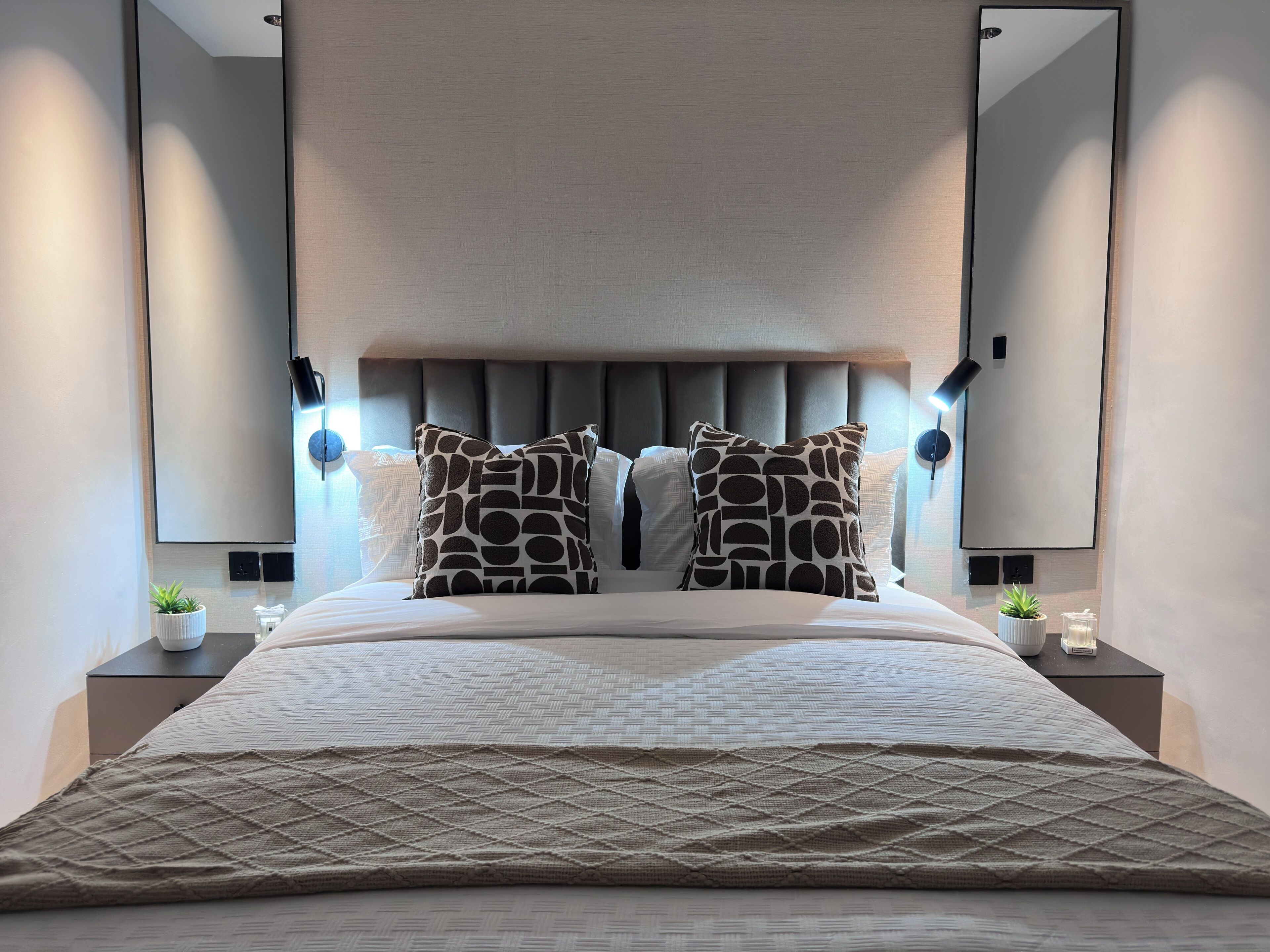 A modern hotel room with a large bed, two decorative pillows, a headboard, bedside lamps, and a small plant on the nightstand.