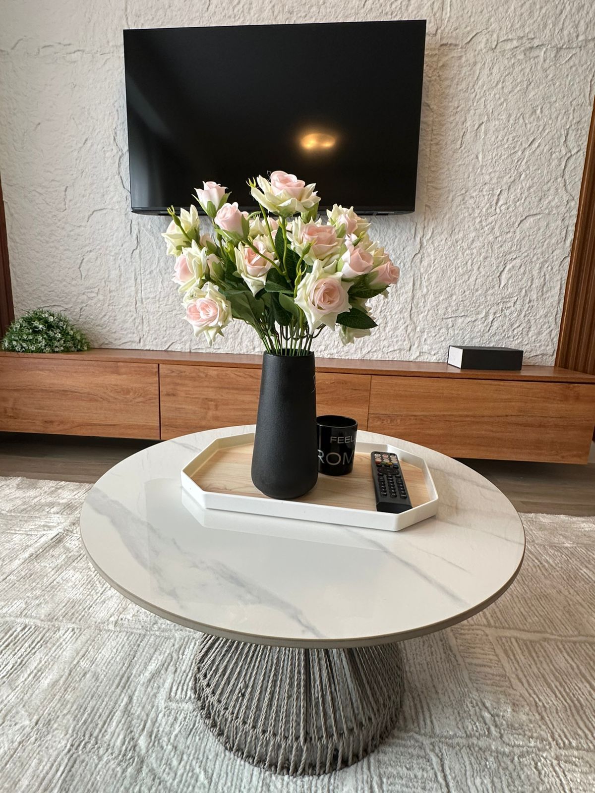 Une table basse ronde en marbre, sur laquelle se trouvent un vase noir contenant des fleurs roses et blanches, une télécommande et une tasse posée sur un plateau.