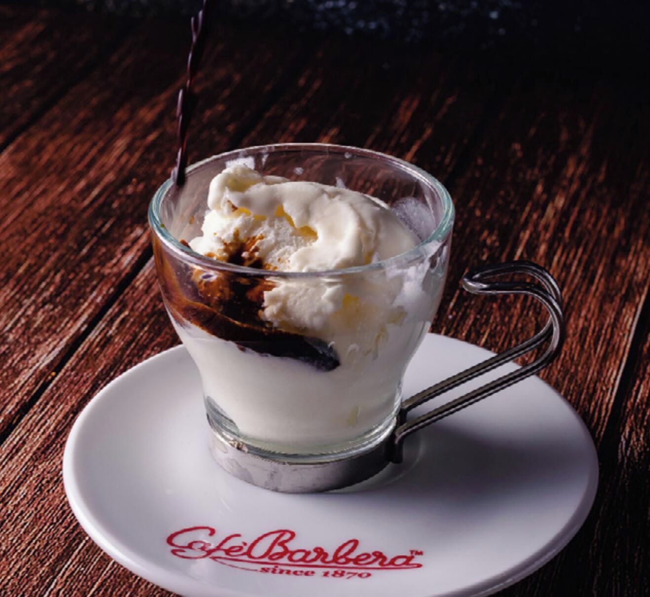 Un postre en una copa de vidrio sobre un plato, con una cuchara, colocado sobre una mesa de madera.