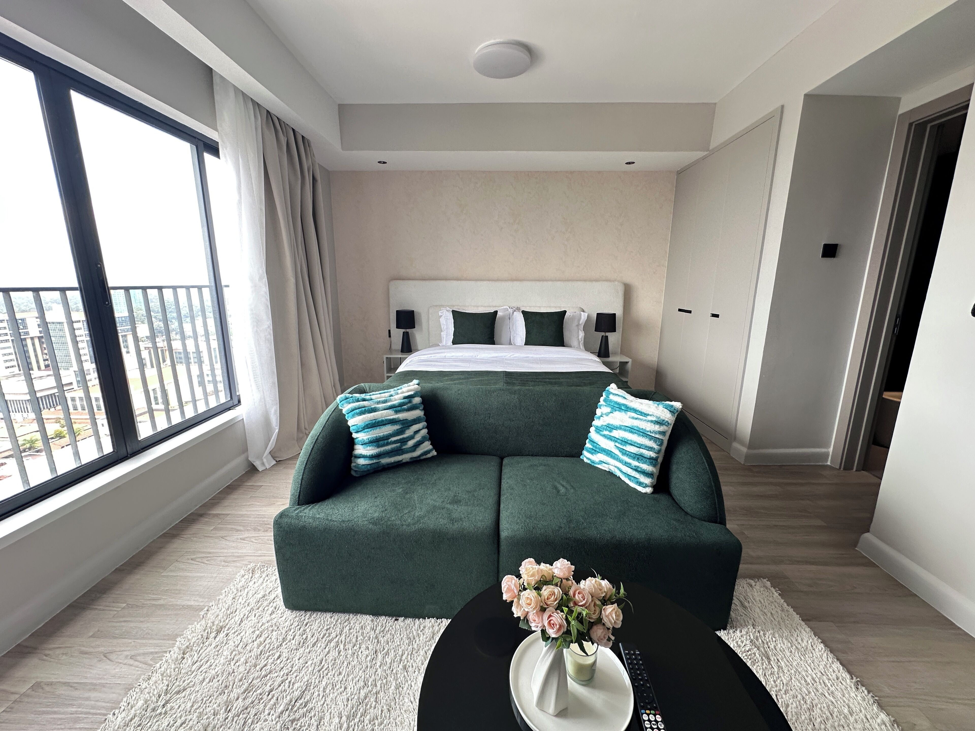 A modern hotel room with a large bed, a green sofa, a white rug, and a vase of flowers.