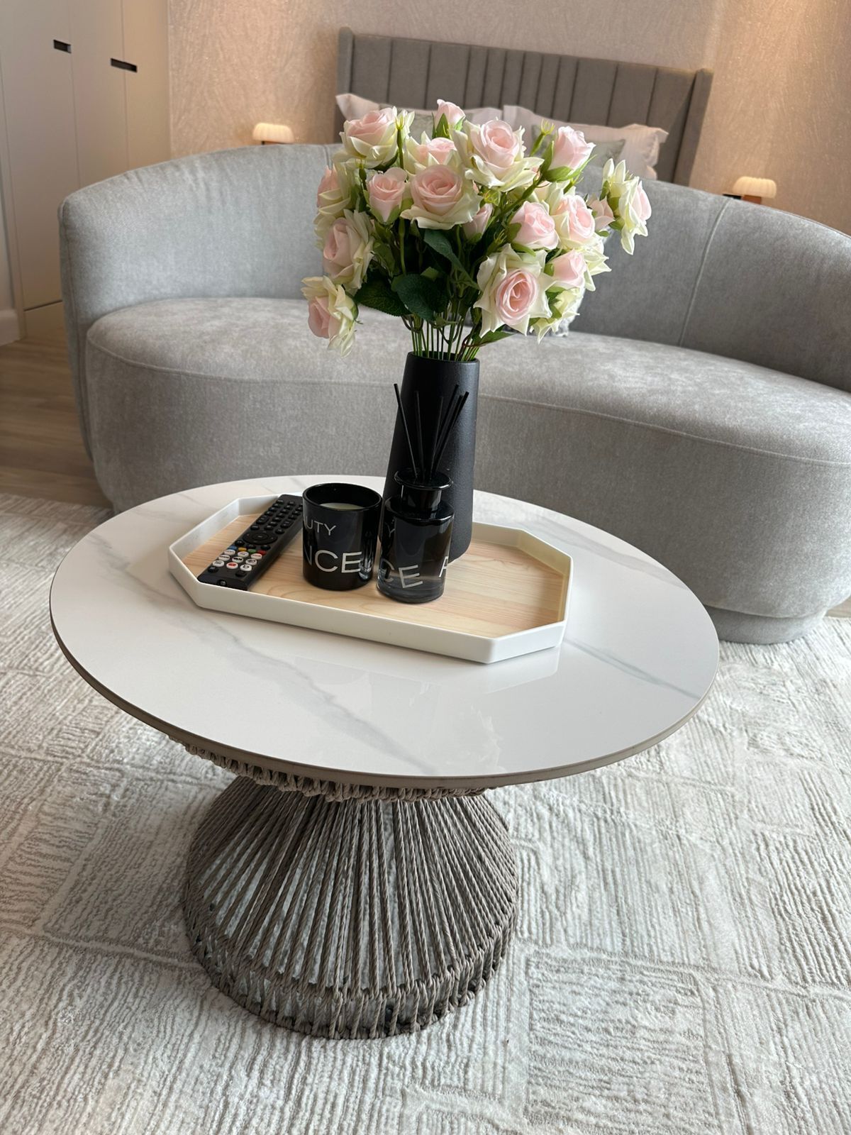 A round coffee table with a tray holding a remote control, two black candles, and a vase of pink roses.