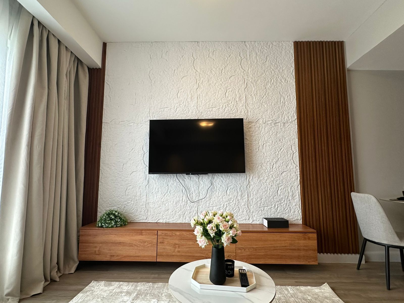 Una sala moderna con un televisor de pantalla plana montado en una pared texturizada, un mueble de madera debajo y una mesa de centro redonda con un jarrón de flores.