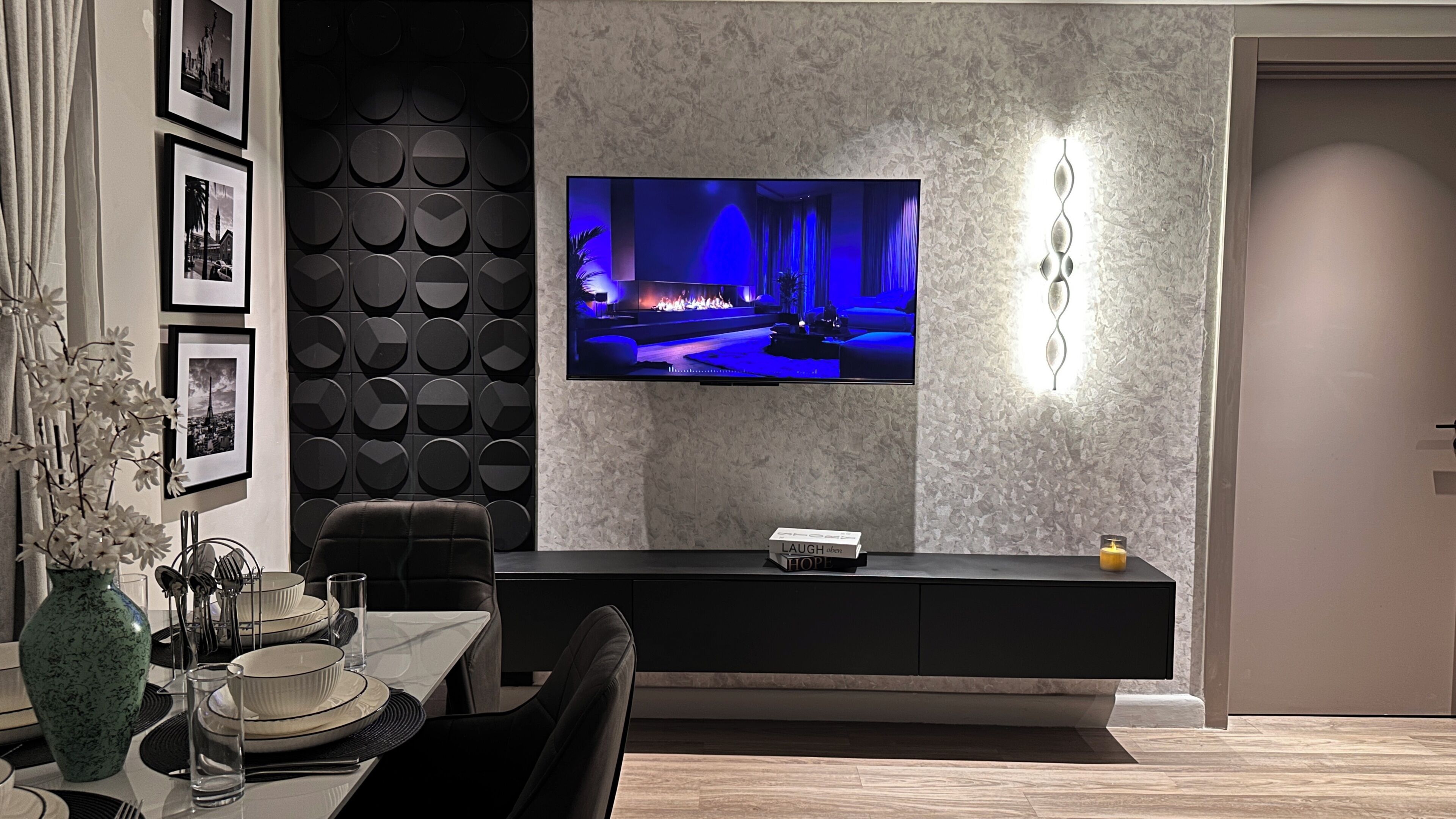 A modern living room with a flat-screen TV mounted on a textured wall, a dark entertainment unit, and a dining area with a vase of flowers.
