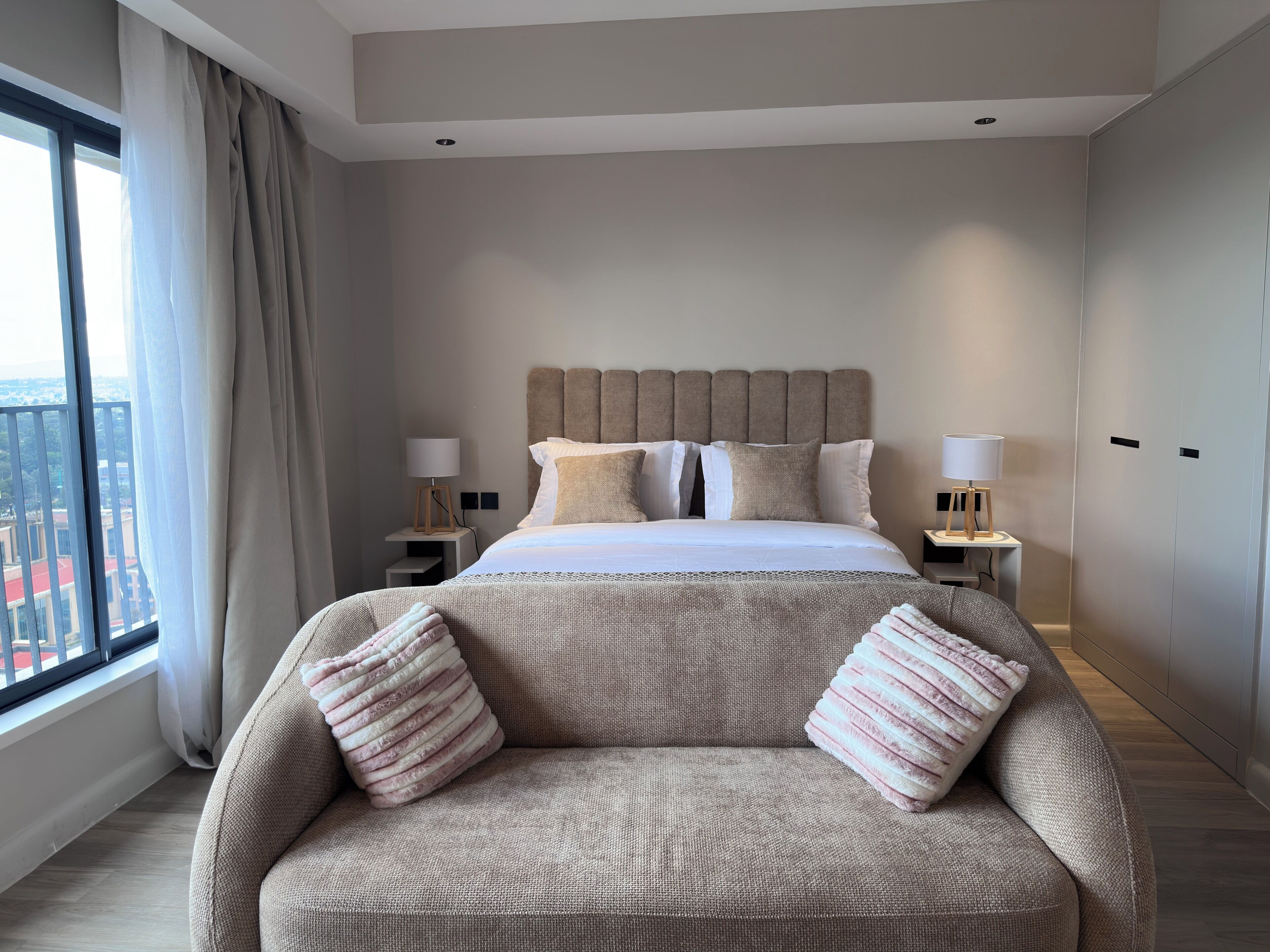 A hotel room with a large bed, two bedside tables with lamps, a sofa with cushions, a window with curtains, and a view of buildings.