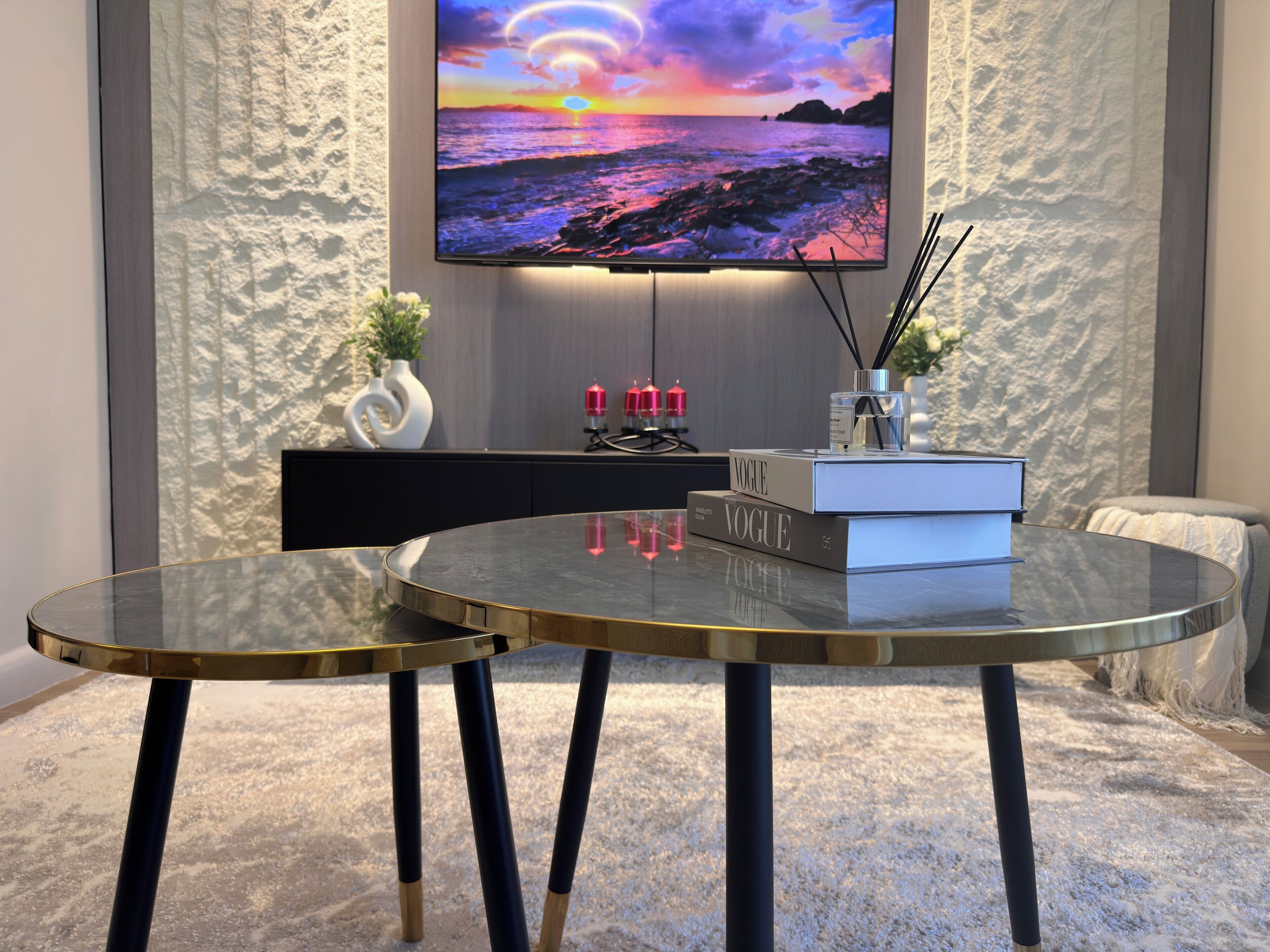 A modern living room with a round glass table, a mounted TV displaying a sunset scene, a decorative vase, and a book titled 'VOGUE'.