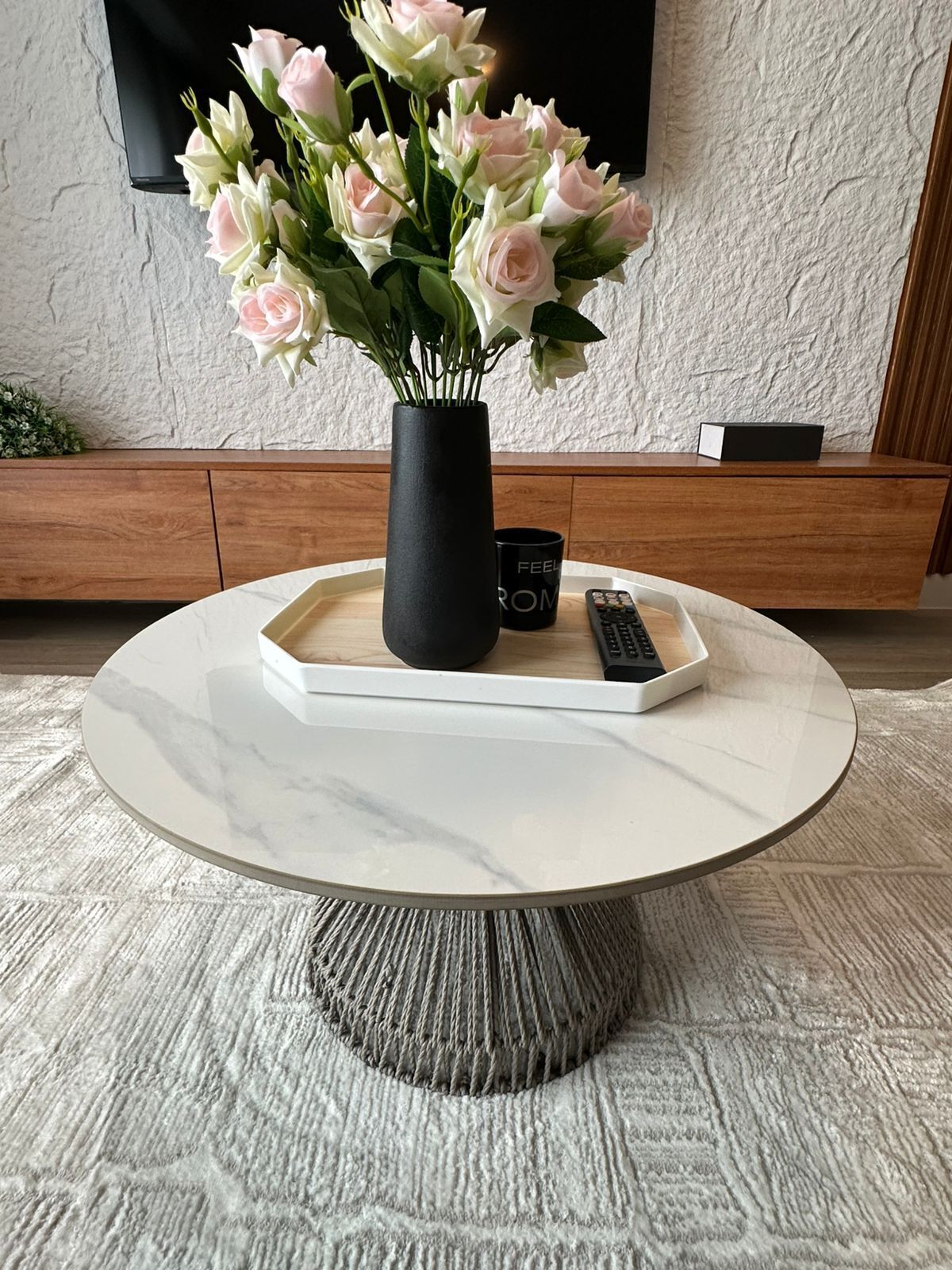 Una mesa redonda con un jarrón de flores, un control remoto y una bandeja con una taza.