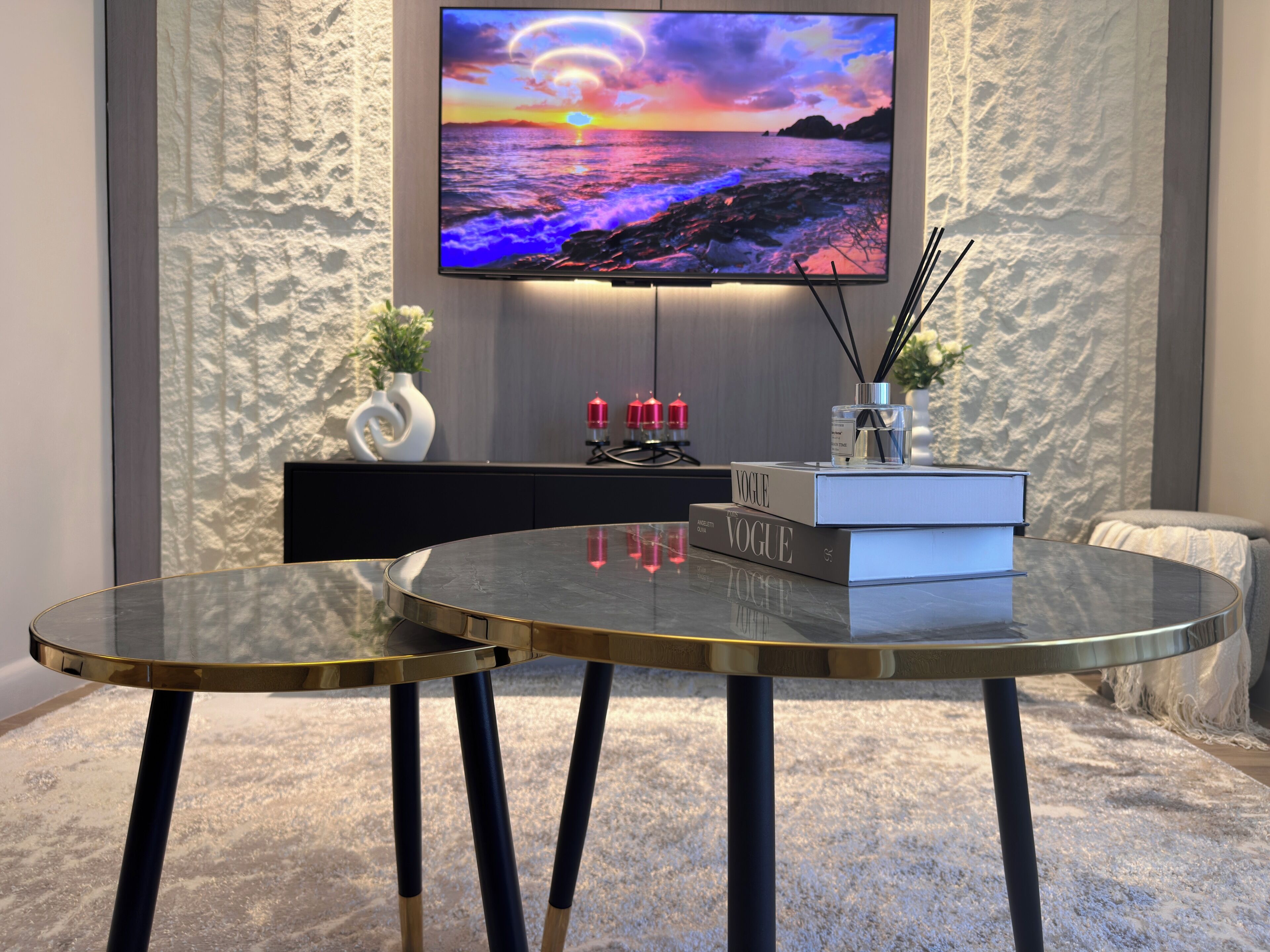 Un salon moderne avec un téléviseur à écran plat, deux tables en verre, un canapé noir, un vase décoratif et un livre intitulé « Vogue ».